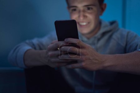 Photo for Cheerful caucasian teenage boy using mobile phone while sitting at night in his room - Royalty Free Image