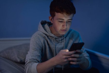Téléchargez les photos : Affectueux caucasien adolescent garçon à l'aide de téléphone mobile tout en étant assis la nuit dans sa chambre - en image libre de droit