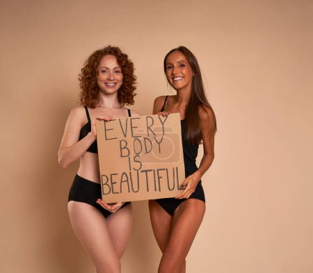 Téléchargez les photos : Deux femmes caucasiennes en sous-vêtements debout sur fond nu en studio et tenant une bannière - en image libre de droit
