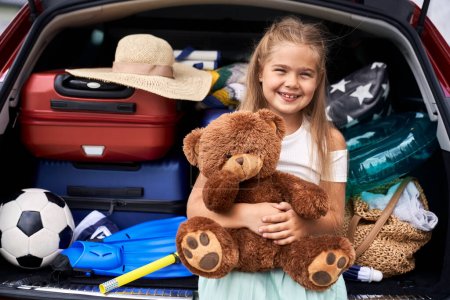 Téléchargez les photos : Portrait de fille caucasienne tenant un ours en peluche et coffre de voiture plein de bagages en arrière-plan - en image libre de droit