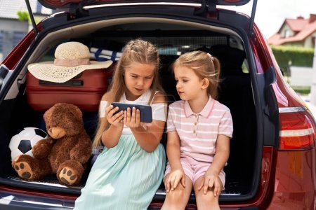 Téléchargez les photos : Deux enfants caucasiens naviguant sur le téléphone portable assis dans le coffre de la voiture - en image libre de droit