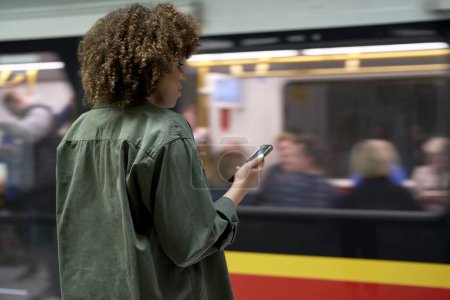 Téléchargez les photos : Jeune femme afro naviguant sur un téléphone portable et sur le point d'arriver au sous-sol - en image libre de droit