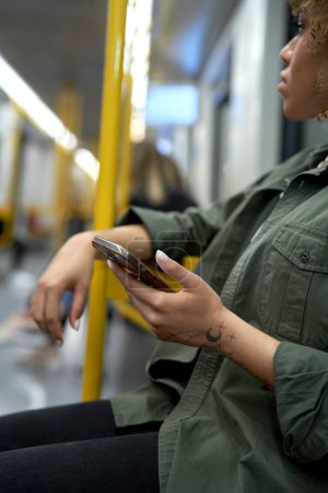 Téléchargez les photos : Image verticale d'une jeune femme afro voyageant en métro et naviguant sur un téléphone portable - en image libre de droit