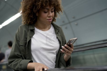 Téléchargez les photos : Jeune femme afro se déplaçant sur l'escalator dans le tube souterrain et textos sur téléphone mobile - en image libre de droit