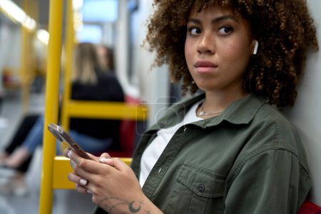Foto de Joven afro mujer navegando por un teléfono móvil en metro y mirando hacia otro lado - Imagen libre de derechos