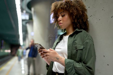 Foto de Joven afro mujer navegando por un teléfono móvil y esperando el metro - Imagen libre de derechos
