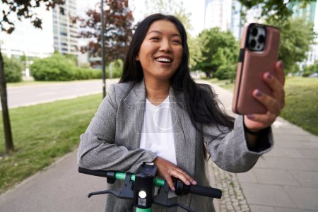 Foto de Mujer china de negocios que tiene videollamada mientras monta un scooter push - Imagen libre de derechos