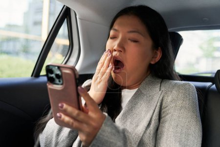 Téléchargez les photos : Femme d'affaires chinoise utilisant le téléphone mobile dans le taxi et le bâillement - en image libre de droit