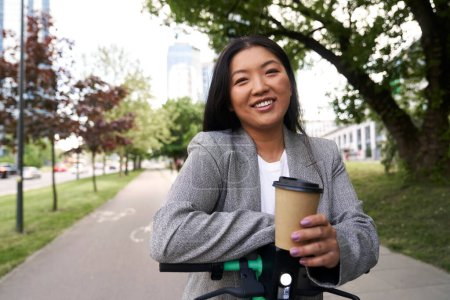 Photo for Portrait of Chinese woman standing on electric scooter in business outfit and holding a cup of coffee - Royalty Free Image