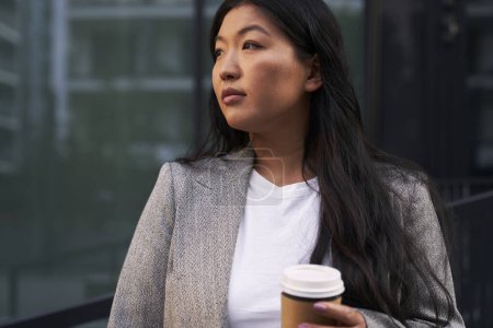 Téléchargez les photos : Femme chinoise d'affaires debout dans la rue avec une tasse de café et regardant loin - en image libre de droit
