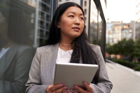 Téléchargez les photos : Femme chinoise d'affaires debout à côté du bâtiment et tenant tablette numérique - en image libre de droit