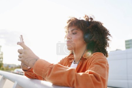 Photo for Black woman wearing headphones and browsing phone while standing on the bridge - Royalty Free Image