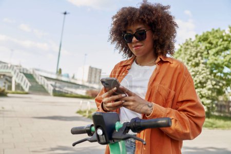 Foto de Mujer negra joven usando teléfono móvil para usar una scooter eléctrica - Imagen libre de derechos