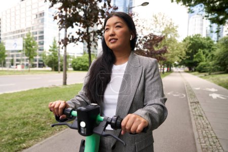 Téléchargez les photos : Femme chinoise chevauchant sur scooter électrique en tenue d'affaires et portant des écouteurs - en image libre de droit