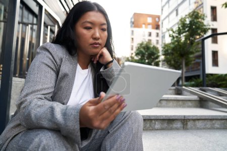 Foto de Mujer china de negocios sentada fuera del edificio y navegando tableta digital - Imagen libre de derechos