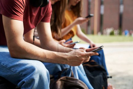 Téléchargez les photos : Étudiants universitaires méconnaissables naviguant sur les téléphones à côté du campus universitaire - en image libre de droit