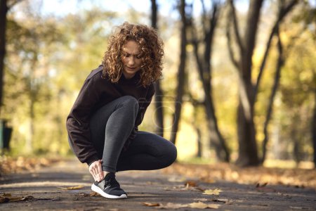 Téléchargez les photos : Femme accroupie et sensation de blessure à la cheville pendant le jogging dans le parc - en image libre de droit