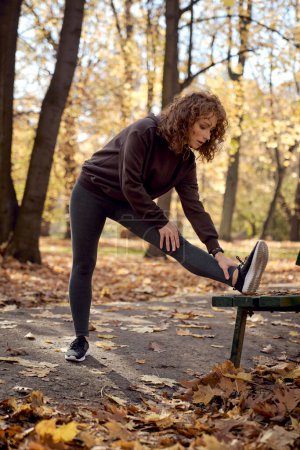 Téléchargez les photos : Pleine longueur de gingembre femme étirement sur banc avant de faire du jogging dans le parc - en image libre de droit