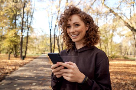 Photo for Caucasian woman browsing mobile phone and looking away in the autumn park - Royalty Free Image