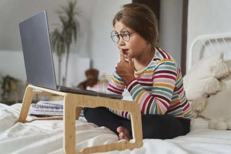 Téléchargez les photos : Caucasienne âge élémentaire fille étudiant sur ordinateur portable tout en étant assis sur le lit - en image libre de droit