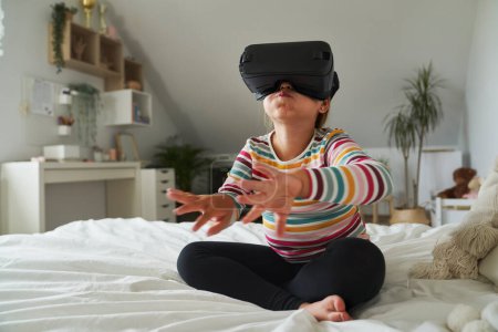 Téléchargez les photos : Age élémentaire fille assise sur le lit et en utilisant des lunettes de réalité virtuelle - en image libre de droit