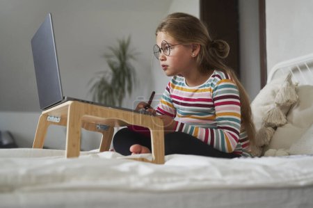 Téléchargez les photos : Caucasienne âge élémentaire fille étudiant sur ordinateur portable tout en étant assis sur le lit - en image libre de droit