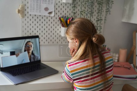 Téléchargez les photos : Caucasienne âge élémentaire fille ayant une conversation vidéo sur ordinateur portable tout en étant assis sur le lit - en image libre de droit