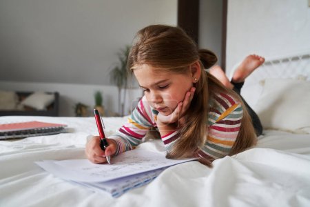 Photo for Elementary age girl studying while lying on front on the bed - Royalty Free Image