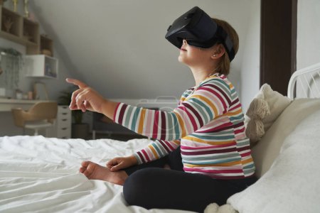 Téléchargez les photos : Age élémentaire fille assise sur le lit et en utilisant des lunettes de réalité virtuelle - en image libre de droit