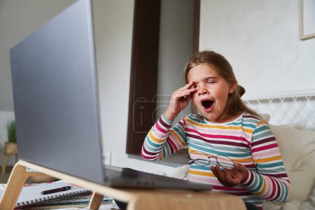 Téléchargez les photos : Caucasien élémentaire âge fille bâiller tout en étudiant sur ordinateur portable - en image libre de droit