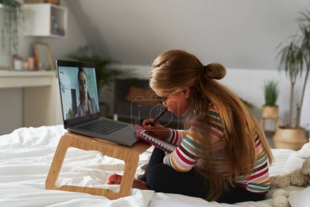 Téléchargez les photos : Caucasienne âge élémentaire fille ayant une conversation vidéo sur ordinateur portable tout en étant assis sur le lit - en image libre de droit