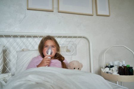 Foto de Niña de edad elemental usando nebulizador en la cama en casa - Imagen libre de derechos