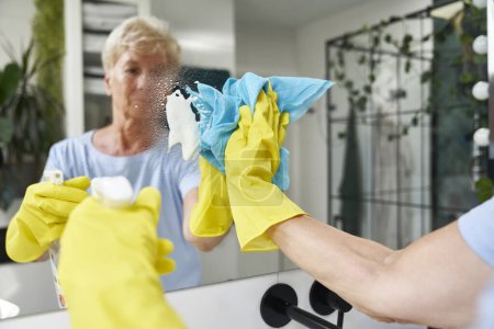 Téléchargez les photos : Senior femme nettoyage miroir dans la salle de bain domestique - en image libre de droit