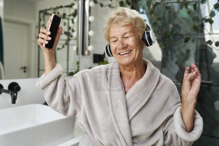Téléchargez les photos : Femme caucasienne senior utilisant des écouteurs et dansant dans la salle de bain - en image libre de droit
