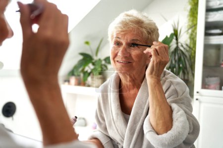 Téléchargez les photos : Femme âgée faisant maquillage des yeux dans la salle de bain - en image libre de droit