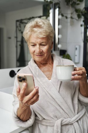 Photo for Senior woman drinking coffee and browsing phone in the bathroom - Royalty Free Image