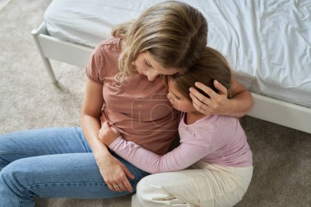 Téléchargez les photos : Mère inquiète embrassant sa triste fille d'âge élémentaire assise sur le sol - en image libre de droit