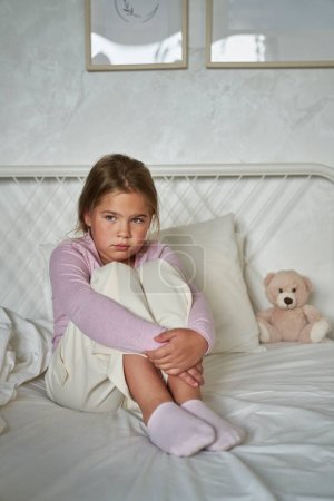 Foto de Triste chica de edad elemental sentado en la cama y mirando hacia otro lado - Imagen libre de derechos