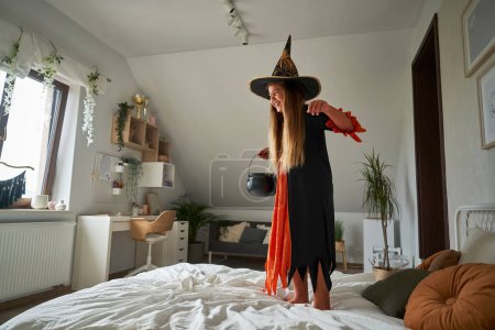 Téléchargez les photos : Caucasienne âge élémentaire fille sautant sur le lit portant costume de sorcière - en image libre de droit
