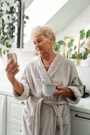 Téléchargez les photos : Femme âgée boire du café et téléphone de navigation dans la salle de bain - en image libre de droit