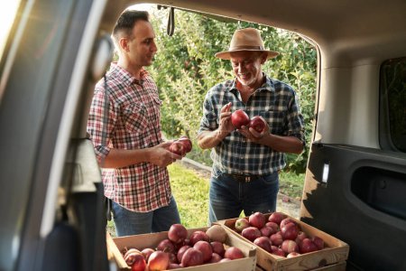 Foto de Dos agricultores de huerta de pie junto a un coche con cajas llenas de manzanas - Imagen libre de derechos
