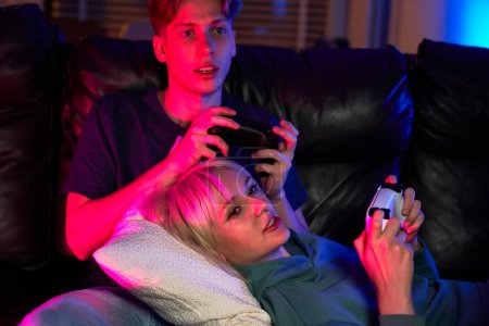Téléchargez les photos : Jeune couple caucasien jouant jeu vidéo avec des tampons de jeu tout en étant assis et couché sur le canapé - en image libre de droit