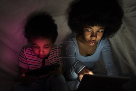 Téléchargez les photos : Deux enfants naviguant tablette numérique sous couverture la nuit - en image libre de droit