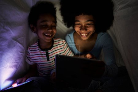 Foto de Dos niños navegando tableta digital en la noche bajo el edredón - Imagen libre de derechos