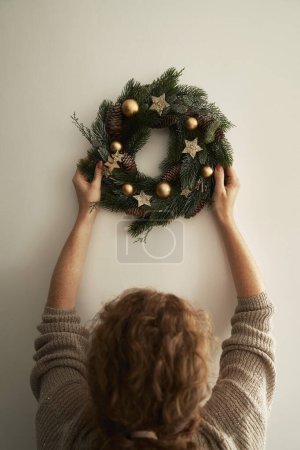 Foto de Imagen vertical de la mujer colgando una corona de Navidad en la pared - Imagen libre de derechos