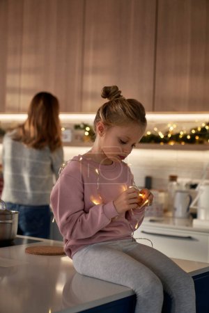 Téléchargez les photos : Fille caucasienne jouer avec des lumières de Noël dans la cuisine - en image libre de droit