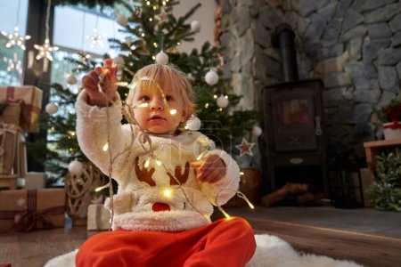 Photo for Toddler sitting on floor and playing with Christmas lights - Royalty Free Image