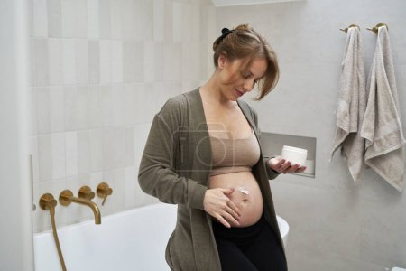 Téléchargez les photos : Femme enceinte caucasienne appliquant hydratant sur son abdomen - en image libre de droit