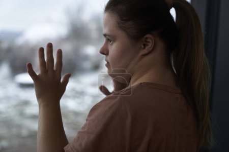Foto de Síndrome de Down pensativo mujer de pie junto a la ventana y mirando hacia otro lado - Imagen libre de derechos