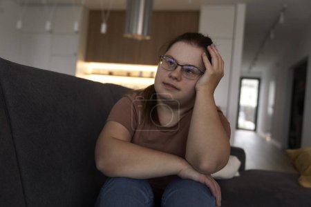Téléchargez les photos : Thoughtful down syndrome femme assise dans le salon et regardant loin - en image libre de droit